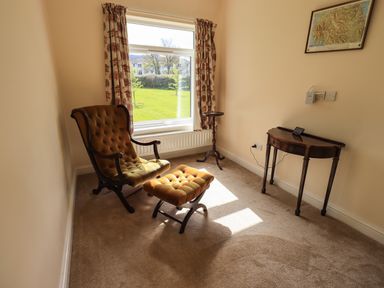 A room with a cushioned armchair and footstool near a window with curtains a small round side table and a wooden console table on carpet at Laal Field House in Keswick