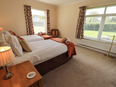 A bedroom with two single beds side by side with pillows and rolled towels on a blanket a wooden bedside table with a lamp two windows with curtains and a chair near one window at Laal Field House in Keswick