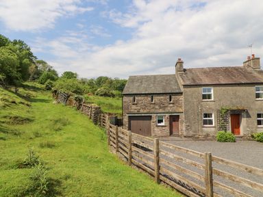 A stone and stucco house with wooden doors next to a grassy hill and wooden fence at Goswick Hall in Bowland Bridge