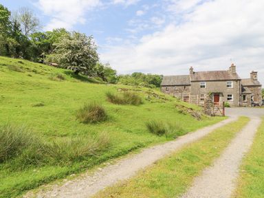 A country house with a gravel driveway and green hillside with trees and bushes at Goswick Hall in Bowland Bridge