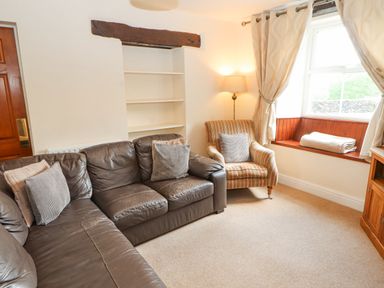 A living room with a leather sofa armchair shelves lamp and window with curtains at Goswick Hall in Bowland Bridge