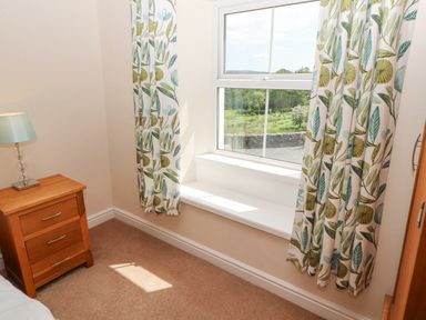A bedroom corner with a wooden bedside table and a lamp by a window with curtains at Goswick Hall in Bowland Bridge