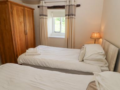 A bedroom with two single beds a wooden wardrobe a window with curtains and a bedside lamp at Goswick Hall in Bowland Bridge