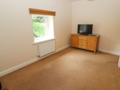 A room with a window and radiator on the left wall and a wooden cabinet with a television and electronic devices on the right side of the room at Goswick Hall in Bowland Bridge