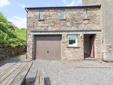 Stone building with garage door and entrance door next to a window and wooden picnic table with bench outside at Goswick Hall in Bowland Bridge