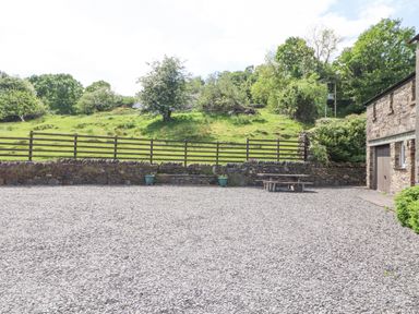 A gravel courtyard with a wooden picnic table and a stone building next to a green grassy hillside at Goswick Hall in Bowland Bridge