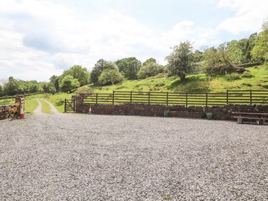 An outdoor gravel area with a gate and wooden fence next to a grassy hillside with trees at Goswick Hall in Bowland Bridge