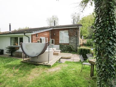 A backyard with a hot tub on a wooden deck chairs and a table next to a house with wood and stone exterior at Lake Lodge in Bowness-On-Windermere