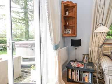 A corner of a living room with a small black table holding books and magazines a black lamp and a silver ice bucket next to a plaid sofa with cream curtains and a wooden corner shelf on the wall at Lake Lodge in Bowness-On-Windermere