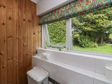 A bathroom with wooden paneling a white toilet three rolls of toilet paper and a window with floral curtains showing a garden outside at Lake Lodge in Bowness-On-Windermere