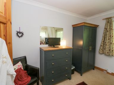 A bedroom corner with a black armchair, a dark dresser with a small TV, a tall dark wardrobe, and floral curtains at Lake Lodge in Bowness-On-Windermere