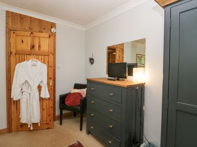 A bedroom corner with a wooden door with a white robe hanging on it a black chair with cushions a dark dresser with a small TV and lamp on top and a dark wardrobe at Lake Lodge in Bowness-On-Windermere