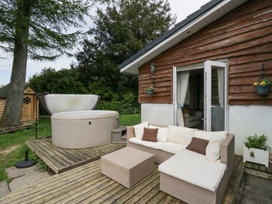 An outdoor patio with wicker sofa and ottoman next to a round hot tub by a wooden house at Lake Lodge in Bowness-On-Windermere