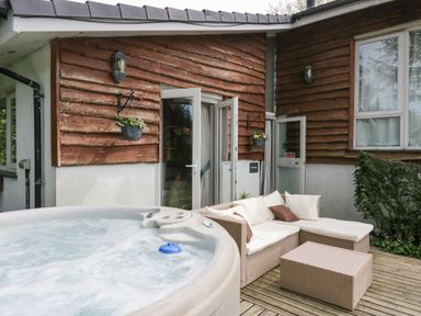 An outdoor patio area with a hot tub and cushioned wicker sofa set beside a wooden cabin exterior at Lake Lodge in Bowness-On-Windermere