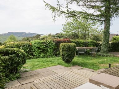 A garden area with a tree, bushes, a wooden deck, a stone patio, and a wooden table with three chairs at Lake Lodge in Bowness-On-Windermere