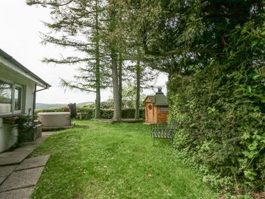 A grassy backyard with trees a wooden shed a metal bench and a hot tub beside a house at Lake Lodge in Bowness-On-Windermere