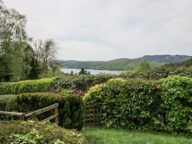 A garden with various green bushes and a wooden fence with a lake and hills in the background at Lake Lodge in Bowness-On-Windermere