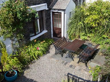 A garden patio with wooden table and benches surrounded by plants and a stone house at Honeysuckle Cottage in Bowness
