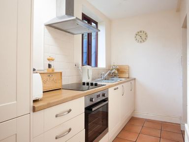 A kitchen with wooden countertop white cabinets an oven a stove a kettle a bread box and a wall clock at Honeysuckle Cottage in Bowness