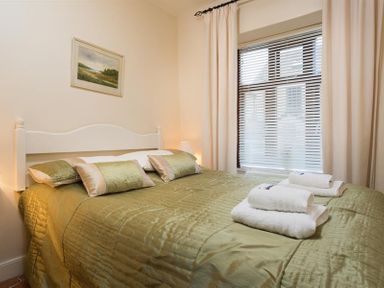 A bedroom with a white headboard bed covered with green bedding and pillows two sets of folded towels on the bed and a window with blinds and curtains at Honeysuckle Cottage in Bowness