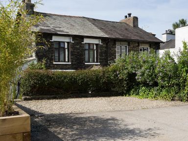 A gravel driveway with plants and bushes in front of a stone house at Honeysuckle Cottage in Bowness