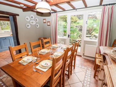 A dining room with a wooden table set for six near windows with curtains at Gunpowder Cottage in Langdale