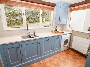 A kitchen with blue cabinets a stainless steel sink a washing machine and a window showing stone wall at Gunpowder Cottage in Langdale
