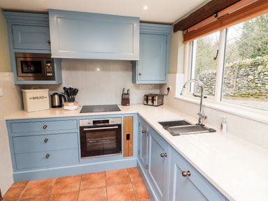 A kitchen with blue cabinets and drawers a microwave oven kettle toaster bread box and a window above the sink at Gunpowder Cottage in Langdale