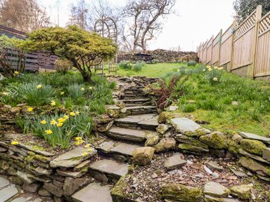 A stone pathway with steps leading up a garden with grass flowers trees and wooden fences at Gunpowder Cottage in Langdale