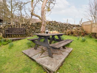 An outdoor garden area with a wooden picnic table on a stone slab and a wooden bench near a stone wall at Gunpowder Cottage in Langdale