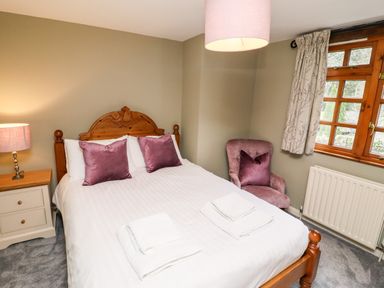 A bedroom with a wooden bed with white linens and purple pillows a bedside table with a lamp a purple armchair near a window with curtains at Gunpowder Cottage in Langdale