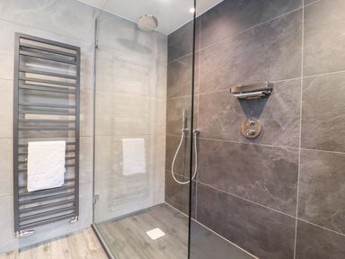 A shower area with a glass partition a towel rack with a white towel and grey wall tiles at Gunpowder Cottage in Langdale