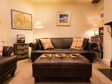 A living room with leather sofa and armchair with plaid pillows a large ottoman with trays and remote controls lamps framed pictures and a bookshelf at Orrest Range in Windermere