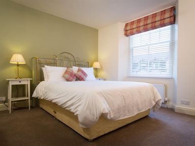 A bedroom with a bed with white bedding and two plaid pillows two bedside tables with lamps and a window with plaid blinds at Orrest Range in Windermere