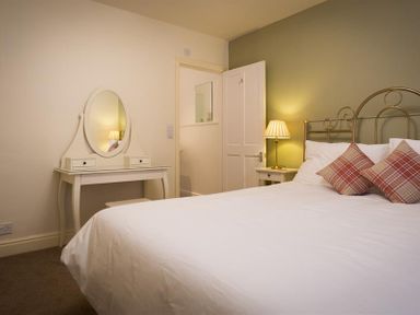 A bedroom with a bed with plaid pillows beside a small nightstand with a lamp and a dressing table with an oval mirror at Orrest Range in Windermere