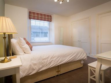 A bedroom with a bed white bedding pillows a lamp on a small table window with blinds and plaid valance two closed doors and a small desk with chair at Orrest Range in Windermere