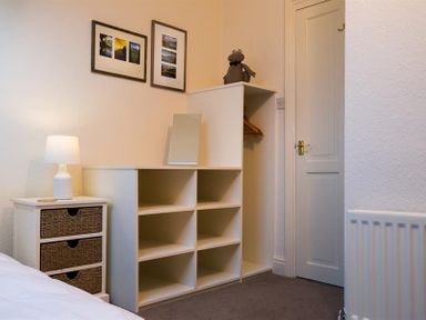 A bedroom corner with a white nightstand with a lamp and wicker drawers a white shelving unit with cubby holes a mirror a stuffed toy and a closed door at Orrest Range in Windermere