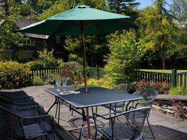 An outdoor patio with a table, chairs, a green umbrella, and a bottle with glasses surrounded by greenery and a house in the background at The Old Barn in Ambleside