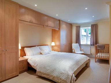 A bedroom with a double bed two bedside lamps built in wardrobes a white chair and a wooden crib at The Old Barn in Ambleside