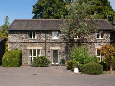 A stone house with four windows a door and various bushes and trees in front at The Old Barn in Ambleside