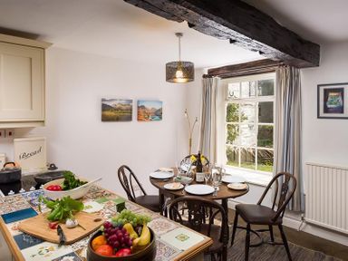 A kitchen and dining area with a round table set for four near a window and a countertop with fruit and vegetables at Camellia Cottage in Bowness