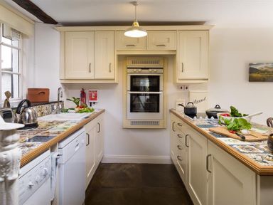 A kitchen with cream cabinets, tiled countertops, a double oven, a kettle, cooking pots, and vegetables on the counter at Camellia Cottage in Bowness