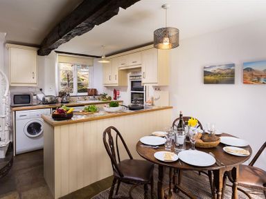 A kitchen with a round wooden dining table set for four and a kitchen island with appliances and food at Camellia Cottage in Bowness