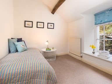 A bedroom with a single bed with striped bedding and pillows a round side table with a lamp and books a window with a blue roller blind and yellow flowers on the windowsill at Camellia Cottage in Bowness