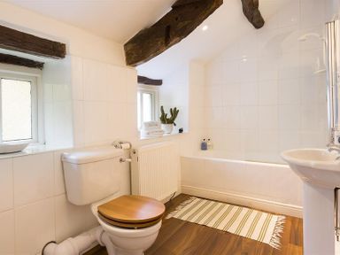 A bathroom with a toilet with wooden seat a bathtub a sink and exposed wooden beams at Camellia Cottage in Bowness