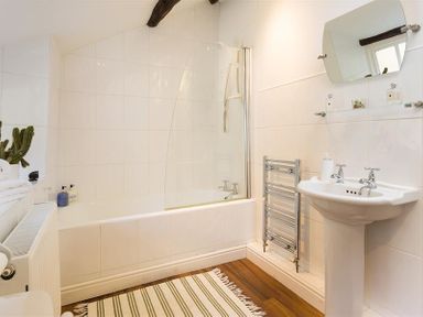 A bathroom with a bathtub shower combination a white sink with a mirror and a striped rug on wooden floor at Camellia Cottage in Bowness