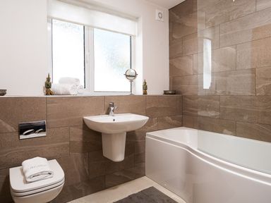 A bathroom with a wall-mounted toilet a pedestal sink a bathtub with glass panel and brown tiled walls at Bowderstone in Keswick