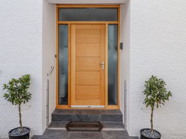 A wooden front door with frosted glass panels on a white textured wall with two potted plants on either side and a welcome mat at Bowderstone in Keswick