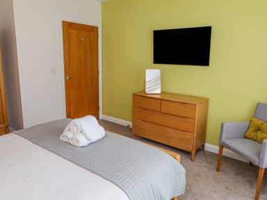 A bedroom with a bed folded towels on it a wooden dresser with a mirror a wall-mounted TV a wooden door and a chair with a cushion at Bowderstone in Keswick
