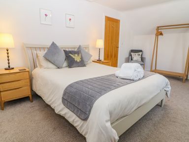 A bedroom with a bed gray pillows and folded towels two wooden nightstands with lamps a wooden door a gray armchair and a wooden clothes rack at Bowderstone in Keswick